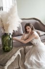 From above magnificent calm woman in elegant white bridal dress touching hair and looking at camera while sitting on floor near elegant sofa in vintage style — Stock Photo