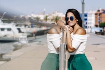 Seitenansicht einer verträumten jungen Frau mit Sonnenbrille, die am Zaun lehnt und am Hafendock wegschaut — Stockfoto