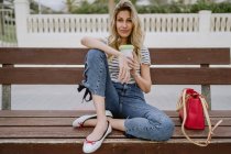 Frau mit Kaffeetasse zum Mitnehmen sitzt an einem Sommertag auf einer Bank am Meer — Stockfoto