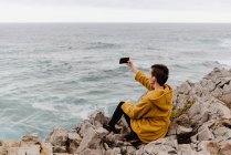 Short haired woman in yellow sweatshirt sitting on rocky seashore and taking selfie on mobile phone on gray cloudy day — Stock Photo