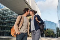 Внизу счастливая пара коллег целуют друг друга, стоя перед современным зданием на городской улице после работы — стоковое фото
