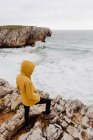 Back view of traveler in yellow warm hoodie standing alone on rocky shore looking at foamy waves on cloudy day — Stock Photo