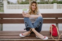Cheerful casual woman with takeaway cup of coffee sitting on city bench at seafront on summer day — Stock Photo