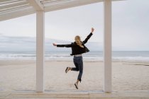 Rückansicht einer trendigen Frau in schwarzer Jacke, die fröhlich mit erhobenen Armen auf einem weißen Holzsteg mit Ozean auf dem Hintergrund springt — Stockfoto