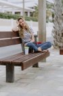 Вид збоку весела жінка сидить на міській лавці на набережній в літній день, дивлячись на камеру — стокове фото