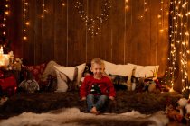 Adorable niño sentado en la habitación llena de decoración de Navidad y mirando en la cámara - foto de stock