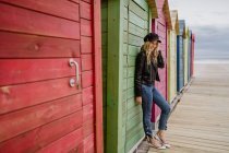 Trendy woman in black cap and leather jacket smiling brightly at camera and leaning on wall of wooden beach cabin — Stock Photo