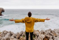 Back view of traveler in yellow warm hoodie standing alone on rocky shore looking at foamy waves on cloudy day raising arms — Stock Photo