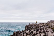 Vista laterale di persona in felpa gialla con cappuccio che calpesta la riva pietrosa circondata da onde schiumose nella giornata grigia nuvolosa — Foto stock