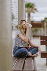 Вид сбоку на веселую случайную женщину, пьющую из чашки кофе, сидящую на городской скамейке у моря в летний день — стоковое фото