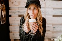 Довгошерста модна жінка тримає келих смачної пінистої кави, облизуючи губи в кафе — стокове фото