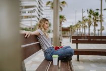 Vue latérale de femme occasionnelle joyeuse avec tasse de café à emporter assis sur le banc de la ville en bord de mer le jour de l'été — Photo de stock