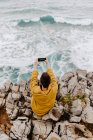 Back view of woman in yellow sweatshirt sitting on rocky seashore and taking selfie on mobile phone on gray cloudy day — Stock Photo