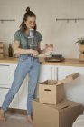 Босоногая женщина в повседневной одежде держит металлические контейнеры и вынимает стеклянную банку из коробок на кухне — стоковое фото