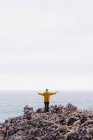 Back view of woman in yellow hoodie stepping on stony shore surrounded by foamy waves on gray cloudy day — Stock Photo