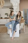 Cheerful casual women laughing and sitting on white washing machine in spacious kitchen having fun at home — Stock Photo
