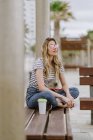 Cheerful casual woman sitting on city bench at seafront on summer day looking away — Stock Photo