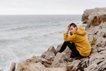 Side view of woman in yellow warm hoodie sitting alone on rocky shore waves on cloudy day and looking at camera — Stock Photo