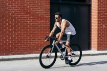 Cycliste homme moderne en vêtements de sport et lunettes de soleil chevauchant un vélo près du mur de briques rouges — Photo de stock