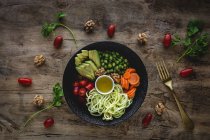 Zucchine crude vegane con piselli, pomodorini, avocado, carote, noci e olio d'oliva in ciotola servite su fondo di legno — Foto stock