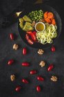 Macarrão vegan cru de abobrinha com ervilhas, tomates cereja, abacate, cenouras, nozes e azeite em tigela servida em fundo escuro — Fotografia de Stock