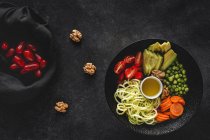 Zucchini raw vegan pasta with peas, cherry tomatoes, avocado, carrots,nuts and olive oil in bowl served on dark background — Stock Photo