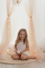 Petite fille tendre en robe blanche assise sur un tapis avec jouet, souriant et regardant à la caméra par des lumières de fées et des draperies élégantes — Photo de stock
