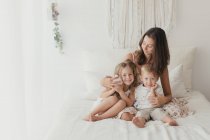 Positiva joven morena sentada en la cama y abrazando a los pequeños niños masculinos y femeninos en el elegante dormitorio - foto de stock