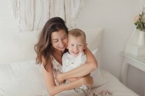 Zufriedene Brünette im weißen Kleid hat Spaß mit glücklichem männlichen Kleinkind, während sie sich auf dem Bett umarmt — Stockfoto