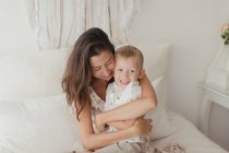 Soddisfatto bruna in abito bianco divertirsi con felice bambino maschio mentre abbraccia sul letto — Foto stock