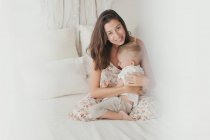 Задоволена брюнетка в білій сукні розважається з щасливим малюком-чоловіком, приймаючи на ліжко — стокове фото
