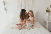 Felice bruna madre divertirsi con bionda carina figlia mentre abbraccia sul letto — Foto stock