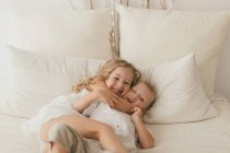 Маленька дівчинка в білій сукні, що обіймає чоловіка веселого молодшого брата на ліжку в стильній кімнаті — стокове фото