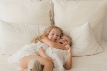 Little girl in white dress embracing male cheerful toddler brother on bed in stylish room — Stock Photo