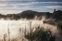 Mysteriöser nebliger See bei holziger Brücke mit großen Bergen und blauem Himmel bei Rototura, Neuseeland — Stockfoto