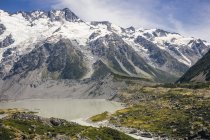 Potenti scogliere ricoperte di vegetazione vicino a un piccolo lago e una grande montagna innevata Cook con skyline blu in Nuova Zelanda — Foto stock