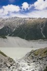 Мощные заросшие скалы возле маленького озера и большая снежная гора Кук с голубым горизонтом в Новой Зеландии — стоковое фото