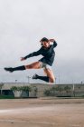 Весела леді-танцівниця в спортивному одязі та кросівках, що розтягують ноги та руку за голову, стрибаючи на відкритому повітрі — стокове фото
