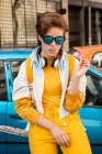 Chica segura en gafas de sol y ropa elegante con auriculares que se extienden goma de mascar contra el coche azul y el edificio moderno - foto de stock