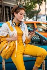Modern teenager with headphones and handbag surfing mobile phone while sitting against colorful cars and modern buildings — Stock Photo