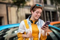 Adolescente moderno con auriculares y bolso de mano que navega por el teléfono móvil mientras se sienta contra coches coloridos y edificios modernos - foto de stock