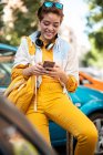 Современный подросток с наушниками и сумочкой для серфинга мобильный телефон, сидя против красочных автомобилей — стоковое фото