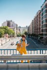 Feminino em roupas brancas e amarelas com mochila em pé na ponte e olhando para o telefone celular contra ruas e edifícios — Fotografia de Stock