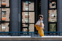 Side view of happy female in headphones and yellow clothes with backpack walking against facade windows with reflection — Stock Photo