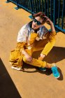 Сверху холодный подросток в солнечных очках слушает музыку в наушниках, сидя на ярком тротуаре с голубым забором — стоковое фото