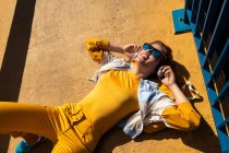 From above chilling teen in sunglasses listening to music with headphones while lying on vivid pavement with blue fence — Stock Photo