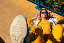 Зверху холодний підліток в сонцезахисних окулярах слухає музику з навушниками, лежачи на яскравій тротуарі з синім парканом — стокове фото