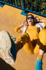 From above chilling teen in sunglasses listening to music with headphones while lying on vivid pavement with blue fence — Stock Photo