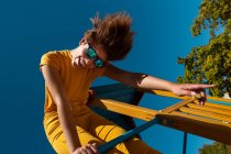 Von unten trendiger Teenager mit Sonnenbrille spielt auf gelbem Querbalken vor strahlend blauem Himmel — Stockfoto