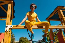 Знизу модний підліток в сонцезахисних окулярах сидить на жовтій поперечині на тлі чистого блакитного неба — стокове фото